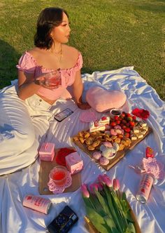 a woman sitting on a blanket in the grass with some food and drinks next to her