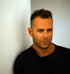 a man in black shirt leaning against wall with his head turned to the side and looking at camera