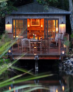 a small cabin is lit up at night on the water's edge with chairs around it