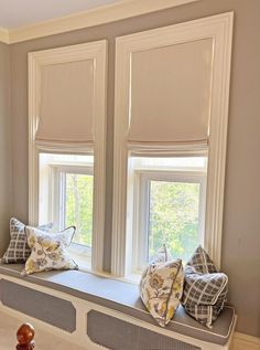 a window seat with pillows on it in front of two windows that have roman shades