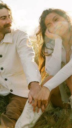 a man and woman holding hands while sitting on the ground in front of some grass