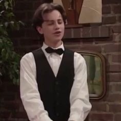 a young man wearing a vest and bow tie standing in front of a brick wall