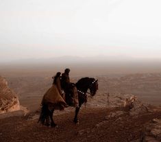 two people riding on the back of a horse near some rocks and desert terrain in the distance