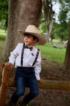 a young boy wearing a cowboy hat and suspenders sitting on a wooden fence in the woods