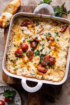 a casserole dish with tomatoes, cheese and bread on a wooden cutting board