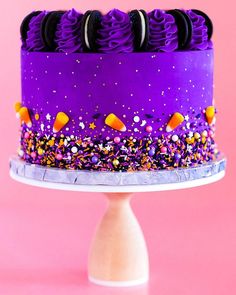 a cake with purple frosting and sprinkles on a white pedestal against a pink background