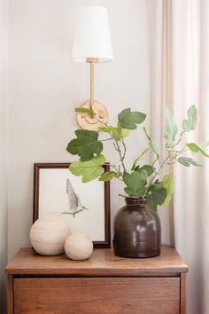 a wooden dresser topped with a vase filled with green leaves next to a white lamp