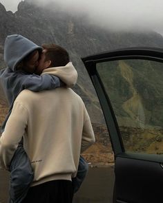 two people embracing each other in front of a car with mountains in the back ground