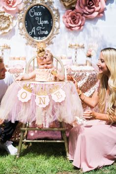 a baby in a pink tutu sits on a chair while two adults sit next to her