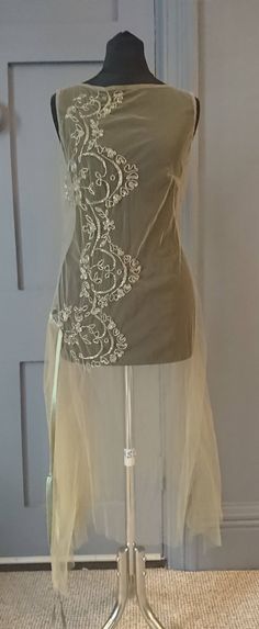 A very pretty 1920s over dress with a wonderful ethereal quality. One piece, a loosely fitted tunic in pale green net, sleeveless, with a high round neck at the front, low at the back. The dress falls to a handkerchief hem, and on the right hip there is a pleated panel, finished at the top with a long green silk ribbon tie / belt. Running down the front on the right side is beaded decoration, stylised deco floral. CONDITION Mostly good, there is one very small hole in the net, and the fabric is Vintage Fashion 1920s, Silk Dancing, Liquid Satin, Beading Netting, Over Dress, Fitted Tunic, Deco Floral, Handkerchief Hem, 1920s Fashion