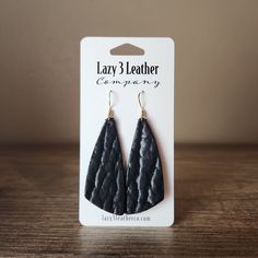 the black leather tear earrings are on display in front of a white card and wooden table