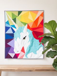 a colorful unicorn quilt hanging on the wall next to a potted plant and wooden bench