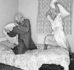 an old man and woman jumping on a bed in a room with two suitcases