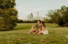 a man and woman are sitting on the grass in front of a cityscape