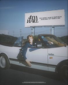 a woman sitting in the driver's seat of a white car next to a billboard