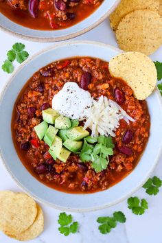 two white bowls filled with chili and cheese next to tortilla chips on the side