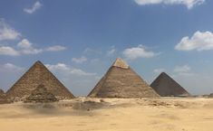 three pyramids in the desert under a blue sky with white clouds and some sand