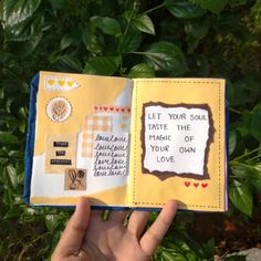 a hand holding an open book in front of some trees and bushes with writing on it