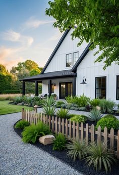 a white house with lots of plants in front of it and a wooden fence around the yard