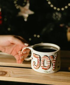 two people holding hands over a coffee cup with the word joy painted on it and christmas lights in the background