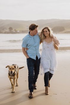 a man and woman walking on the beach with a dog behind them, holding hands