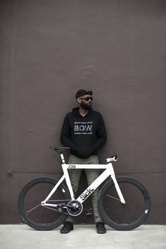 a man standing next to a bike with his hands on the handlebars in front of a gray wall