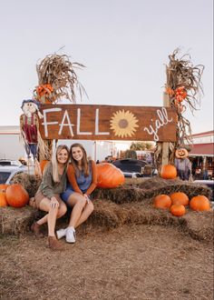 two women sitting on hay bales in front of a fall sign