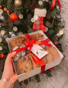 a person holding a box of cookies in front of a christmas tree with presents on it