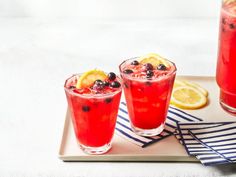 two glasses filled with red liquid and topped with blueberries on a tray next to sliced lemons