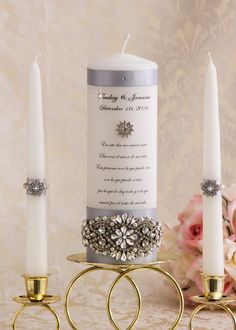 two white candles are sitting next to each other on a table with gold rings and flowers