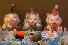 three white cats wearing party hats sitting at a table with cupcakes and cakes