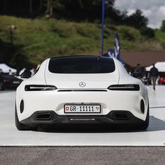 the rear end of a white sports car parked in a parking lot with people walking by