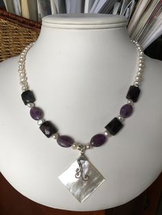 The focal point of this necklace is a rectangular pendant of white Mother of Pearls, which was handcrafted in Bali. The strand combines faceted ovals and faceted of rectangles of amethyst, grey freshwater pearls and silver accents. The necklace is 18 inches long, is strung on jeweler's wire and has a sterling silver lobster clasp. Item number 0519-SN8 White Amethyst Pendant Jewelry, White Amethyst Pendant Necklace, Elegant Rectangular Gemstone Beads Jewelry, Elegant Purple Rectangular Necklace, Faceted Silver Pearl Necklace For Gift, Spiritual Rectangular Gemstone Beads Jewelry, Handmade Purple Rectangular Necklace, Elegant Purple Necklace, Rectangular Purple Amethyst Necklaces