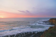 the sun is setting over the ocean with waves crashing on the shore and cliffs in the foreground