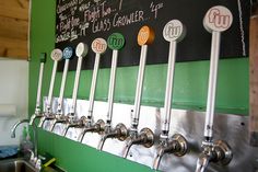 a row of beer taps mounted to the side of a green wall with chalk writing on it