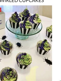 cupcakes decorated with green and purple icing are on a table next to a box of black bugs