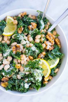 a white bowl filled with greens, chickpeas and lemon wedges on top of a marble table