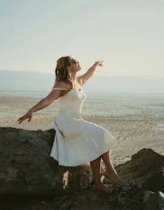 a woman in a white dress is sitting on a rock and pointing to the sky