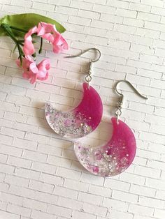 two pink and white moon shaped earrings on a brick wall with flowers in the background