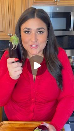 a woman in a red shirt is holding up an eggplant and looking at the camera