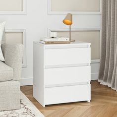 a white night stand with a lamp on it in front of a couch and window