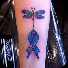 a blue ribbon and dragonfly tattoo on the left arm, with words underneath it