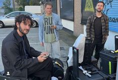 three men standing next to each other with luggage in front of them and one man looking at his cell phone