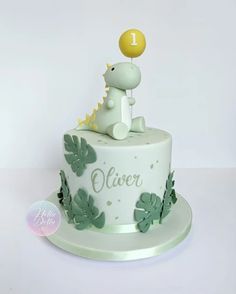 a white cake with green leaves and a dinosaur holding a yellow balloon on it's top