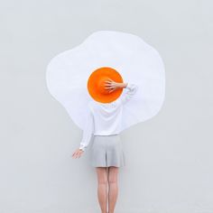 a woman holding an orange frisbee in front of her face while standing against a white wall