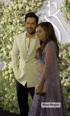 two people standing next to each other in front of a flower covered wall with white flowers