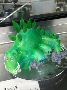 a cake shaped like a green dragon on top of a metal counter with a sign that says don't touch it