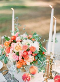 an arrangement of peaches, roses and greenery sits on a table with candles