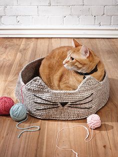 an orange cat laying in a basket with balls of yarn on the floor next to it