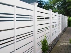 a white privacy fence with plants growing on the side and brick walkway next to it